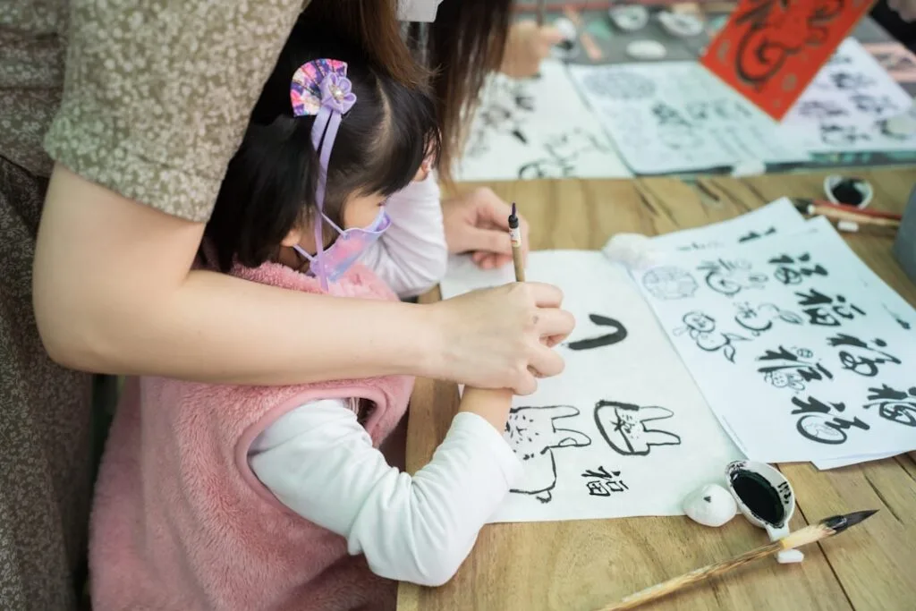Child learning brush painting at a calligraphy workshop, adult guiding.