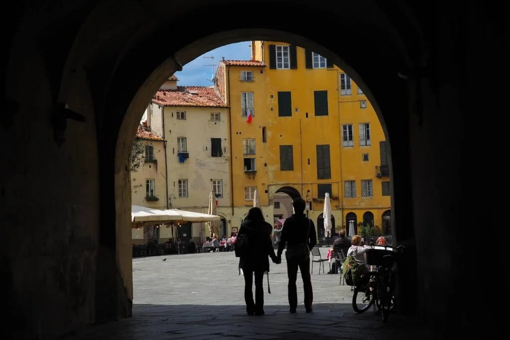 Silhouetted couple holding hands under an arch in Lucca's sunny piazza
