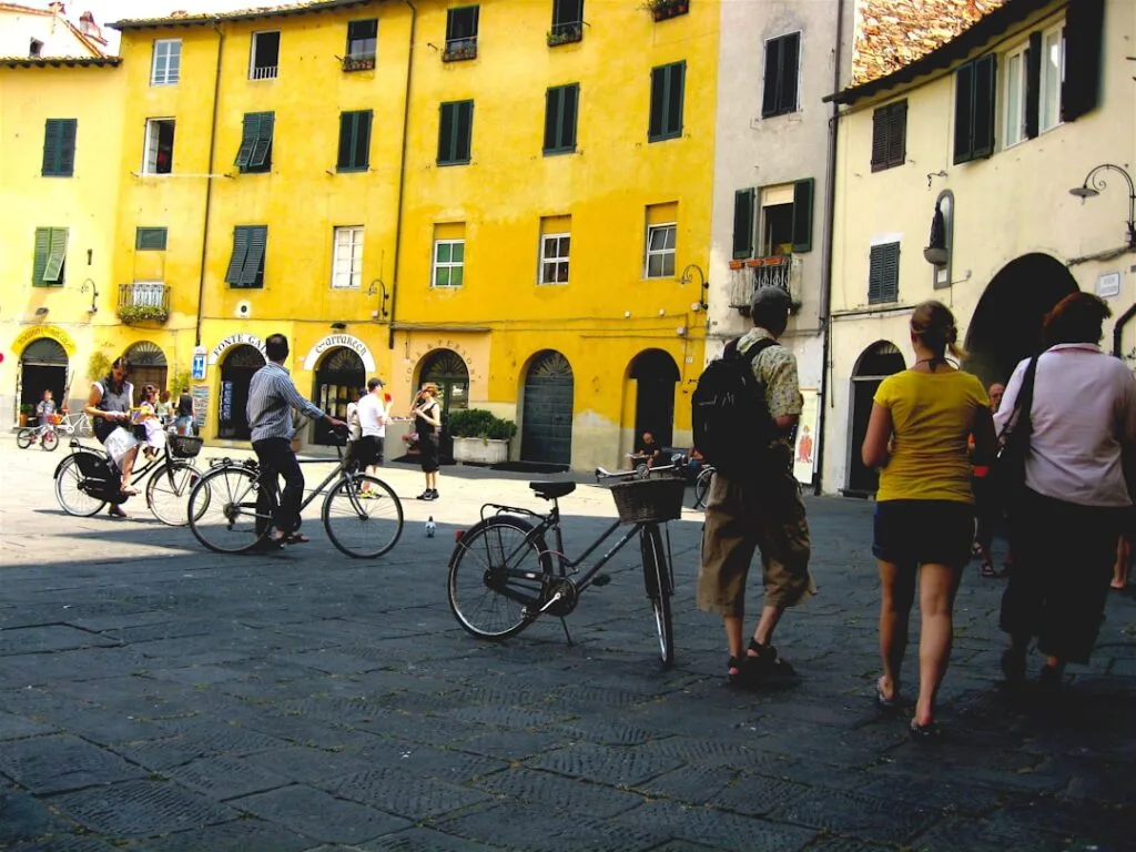 Cyclists and pedestrians in front of yellow buildings at Lucca's Piazza dell'Anfiteatro