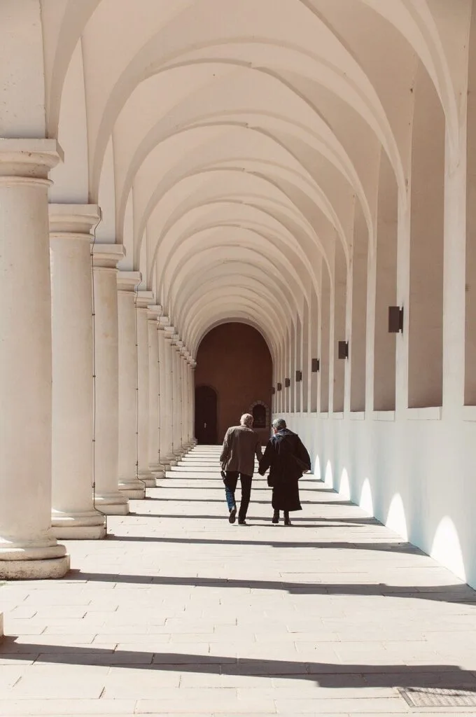 Two people walking under arched cloister in Lucca, columns casting shadows